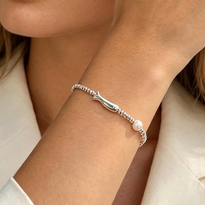 May include: A silver bracelet with a beaded design and a central decorative element. The bracelet features a small, round, white pearl. The woman in the image is wearing a cream-coloured blazer.