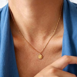 May include: A gold-colored necklace with a sunburst pendant. The pendant is a small, round disc with radiating lines. The necklace is worn against a blue top.