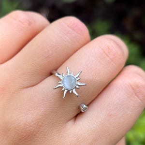 May include: A silver-colored ring featuring a sunburst design with a central, round, pale blue stone. The ring is open and adjustable, with the sun's rays extending outwards. The ring is worn on a finger.