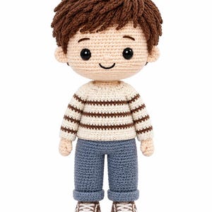 Pode incluir: Uma boneca de crochê feita à mão com cabelo de lã castanho, olhos de contas pretas e um sorriso bordado. A boneca veste uma camisola de manga comprida creme com riscas castanhas horizontais, calças azuis com bainha e sapatilhas castanhas e brancas.