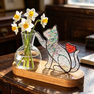 Peut inclure: Un vase en verre transparent contenant des jonquilles blanches et jaunes est posé sur une base en bois, à côté d'une figurine de chat en vitrail avec une queue en forme de cœur rouge. La figurine de chat a un aspect irisé et texturé.