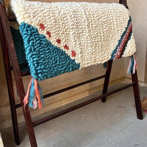 May include: A rectangular rug featuring a thick, cream-colored tufted texture with a large teal triangular section on the left. A diagonal row of small red accents separates the colors, while a striped band of teal, pink, and brown sits on the right.