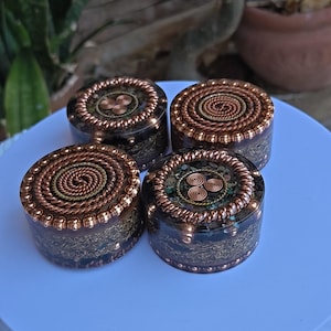 May include: Four round, decorative objects with copper-colored wire detailing. Each has a spiral or swirl design on top, with a dark, patterned base. The objects are displayed on a white surface, with greenery in the background.