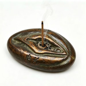 Peut inclure: Porte-encens en bronze avec un bâton d'encens en combustion dégageant de la fumée. Le support ovale présente des détails sculptés et des caractères japonais. L'encens est placé dans une petite dépression circulaire.