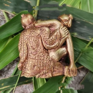May include: A gold-toned figurine depicting two figures in an embrace, with one figure wearing a textured, patterned garment featuring a circular emblem on the back. The sculpture has a metallic finish and shows intricate detailing on the clothing.