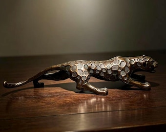 May include: A bronze-toned metal figurine of a leopard in a low, prowling stance. The sculpture features a textured surface with raised, rounded spots across its body, a long extended tail, and a focused head position with an open mouth.
