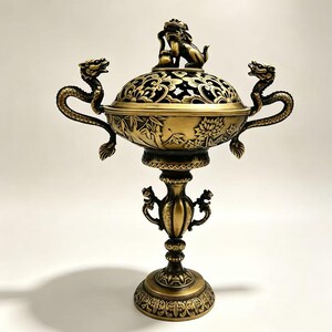 May include: A metallic gold-toned incense burner featuring a lid topped with a mythical creature figurine. The bowl displays embossed floral and bamboo motifs, flanked by two dragon-shaped handles. It stands on a pedestal base with ornate details.