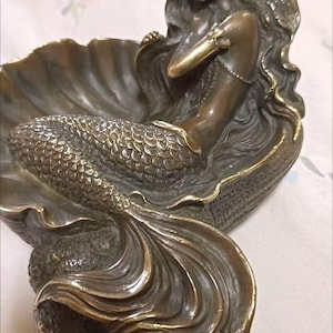 Op de afbeelding: Een bronskleurig beeldhouwwerk van een zeemeermin die rust in een grote, open schelp. De zeemeermin heeft lang, golvend haar en een gedetailleerde, gestructureerde staart. Het beeld heeft een metallic afwerking met accenten op de randen van de schelp en de vorm van de zeemeermin, wat zorgt voor een ruimtelijke uitstraling.