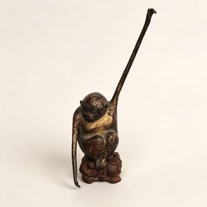 Puede incluir: Una figura de mono de bronce con una pátina marrón oscura y dorada, representada en posición sentada sobre una base texturizada. El mono tiene un brazo extendido hacia arriba y el otro brazo colgando, con rasgos faciales detallados y un acabado desgastado.