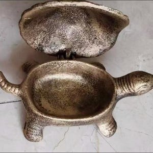 May include: A metallic turtle-shaped trinket box with a textured, antique bronze finish. The hinged shell lid is open, revealing a hollow interior. The turtle has a defined head, four legs, and a tail, with a rough, weathered surface texture throughout.