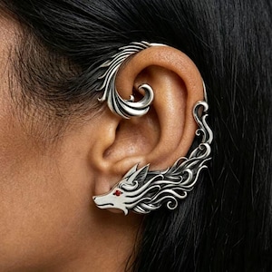 May include: A silver ear cuff featuring a sculpted wolf head with a small red stone eye. The design extends along the outer edge of the ear with intricate, flowing, flame-like metalwork that curls upwards towards the top of the ear cartilage.