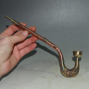 Pode incluir: Um cachimbo de cobre e latão com um design curvo. O cachimbo tem um longo tubo reto com um bocal de latão e uma tigela curva. A tigela e a seção curva do cachimbo são de latão, enquanto o tubo é de cobre.