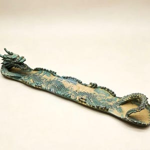 May include: A long, ceramic incense holder in the shape of a dragon. The dragon is teal and has intricate details, with a textured body and a curled tail. The holder is approximately 12 inches long and has a beige interior.