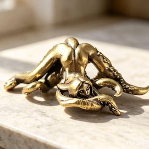 以下が含まれることがあります： 真鍮色の金属製フィギュアで、人間のような胴体と外側に伸びる複数のタコのような触手を持つ生き物を表現しています。表面は金属光沢があり、暗い凹部が手足の吸盤の質感を強調しています。