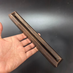Peut inclure: Un objet long et rectangulaire de couleur bronze, doté d'un motif texturé en forme de grille sur ses surfaces extérieures. L'article présente un canal profond et étroit sur toute sa longueur, formant une structure ouverte en forme de gouttière.
