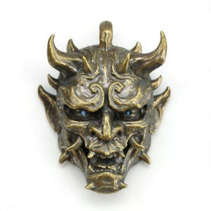 May include: A bronze-colored, detailed mask with horns, sharp teeth, and blue eyes. The mask has an aged appearance, with intricate carvings and a loop at the top for hanging. The overall design suggests a mythical or fantasy theme.