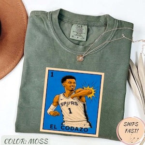 Puede incluir: Una camiseta de color musgo con un gráfico de un jugador de baloncesto con la camiseta de los Spurs. El gráfico es una ilustración estilizada con el texto "EL CODAZO" y el número 1. La camiseta también tiene el texto "COMFORT COLORS" y "S-P-CH".
