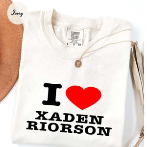 Op de afbeelding: Ivoor kleurig t-shirt met de tekst "I ♥ XADEN RIORSON" in zwart en rood. Een gouden ketting met een hanger is over het shirt gedrapeerd. Het shirt is opgevouwen en heeft een label met "Comfort Colors".