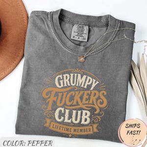 Könnte beinhalten: Ein pfefferfarbenes T-Shirt mit einem Grafikdruck, der "GRUMPY FUCKERS CLUB LIFETIME MEMBER" in beigen und weißen Buchstaben zeigt. Das T-Shirt hat einen Rundhalsausschnitt und ein Comfort Colors Markenetikett im inneren Kragenbereich.