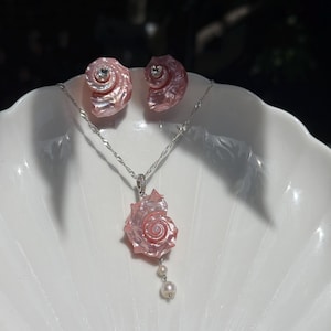 May include: A jewelry set consisting of a silver chain necklace with a pink spiral seashell pendant and two small white pearls, accompanied by a pair of matching pink spiral seashell stud earrings featuring small clear stones at the center.