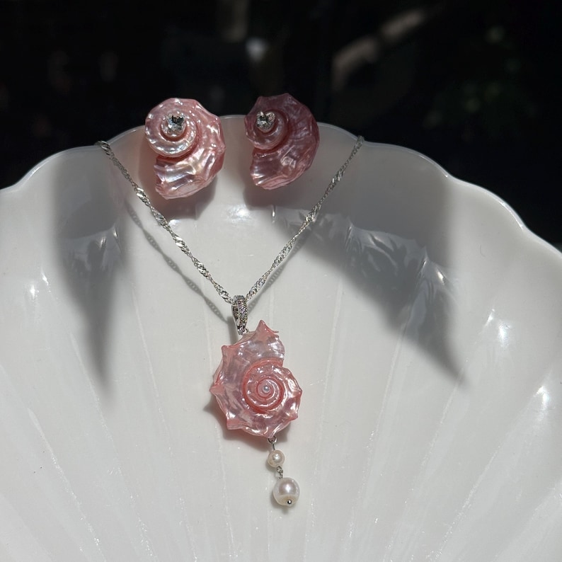 Puede incluir: Un conjunto de joyer&iacute;a que consta de un collar de cadena plateada con un colgante de concha marina rosa en espiral y dos peque&ntilde;as perlas blancas, acompa&ntilde;ado de un par de pendientes de bot&oacute;n a juego con forma de concha rosa y piedras claras.