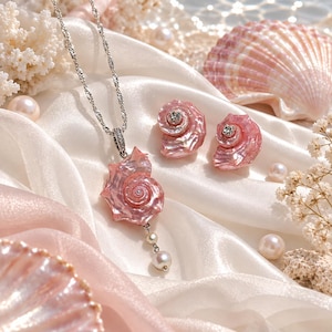 Puede incluir: Un conjunto de joyer&iacute;a que incluye un collar de cadena plateada con un colgante de concha marina rosa iridiscente y dos perlas blancas colgantes, acompa&ntilde;ado de pendientes de bot&oacute;n a juego en forma de concha rosa con una peque&ntilde;a piedra clara.