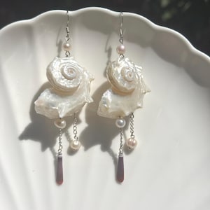 Op de afbeelding: Een paar oorbellen met witte spiraalvormige schelpen, kleine witte parels en zilverkleurige metalen kettinkjes. Elke oorbel heeft een donker roodbruine langwerpige kraal aan het uiteinde van een kettinkje, hangend aan een zilverkleurige haak.