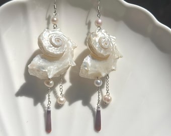 Puede incluir: Un par de pendientes colgantes con caracolas blancas en espiral, pequeñas perlas blancas y cadenas de metal plateado. Cada pendiente incluye una cuenta alargada de color marrón rojizo oscuro al final de una cadena, suspendida de un gancho plateado.
