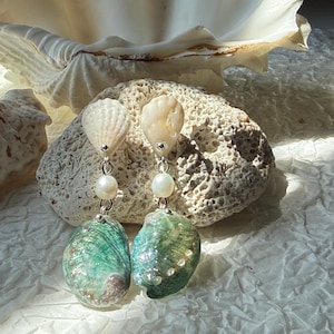 May include: Dangling earrings with natural seashells and pearls. The earrings feature a top shell, a pearl, and a larger, iridescent green and blue shell. The earrings are displayed on a textured, light-coloured stone.