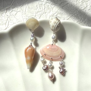 May include: A pair of asymmetrical drop earrings featuring natural seashells and freshwater pearls. One earring has a smooth tan shell and a cone-shaped shell, while the other features a pink fan-shaped shell accented with dangling white and purple pearls.