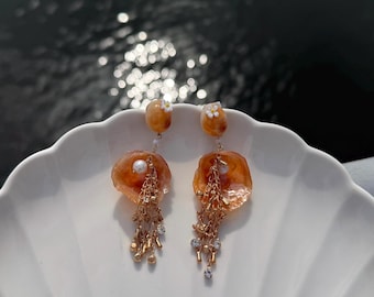 以下が含まれることがあります： オレンジ色の貝殻のようなディスク、小さな白い花のアクセント、そして小さな金属製のビーズと透明なクリスタルが付いたカスケード状のゴールドトーンのチェーンが特徴のダングルイヤリング。白い貝殻型の皿の上に置かれています。