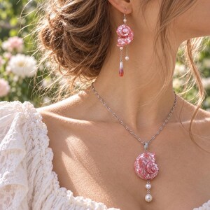 Puede incluir: Un conjunto de joyer&iacute;a que consta de un collar de cadena en tono plateado y un pendiente colgante a juego. Ambas piezas presentan un colgante de concha espiral rosa iridiscente, acentuado con peque&ntilde;as perlas blancas y un dije de gota con punta roja.