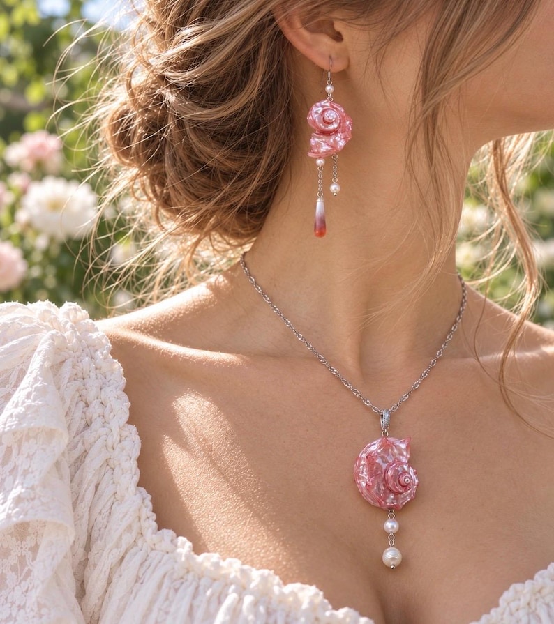 Puede incluir: Un conjunto de joyer&iacute;a que consta de un collar de cadena en tono plateado y un pendiente colgante a juego. Ambas piezas presentan un colgante de concha espiral rosa iridiscente, acentuado con peque&ntilde;as perlas blancas y un dije de gota con punta roja.