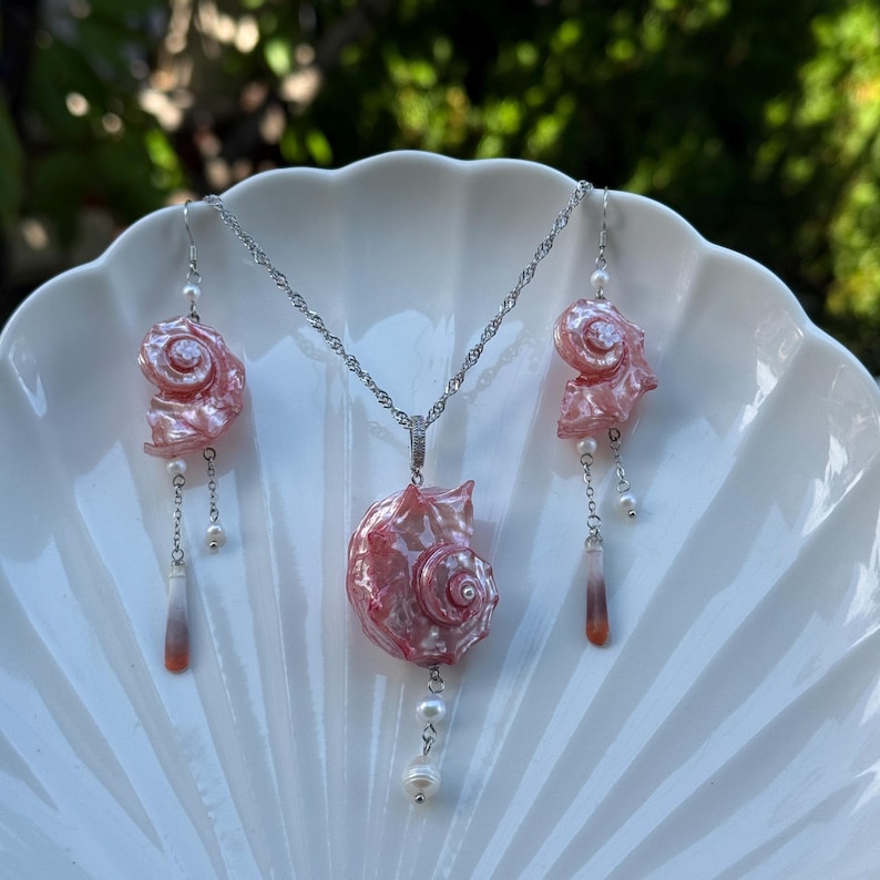Puede incluir: Un conjunto de joyer&iacute;a compuesto por un collar de cadena plateada y dos pendientes colgantes a juego. Cada pieza presenta un dije de concha espiral rosa iridiscente, acentuado con peque&ntilde;as perlas blancas y colgantes delgados, transl&uacute;cidos y degradados.