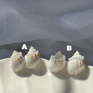 Puede incluir: Cuatro pendientes de concha, dos pares etiquetados A y B. Cada concha es de color blanco y crema, adornada con una hilera de pequeñas perlas blancas en la parte superior. Los pendientes se muestran en un plato blanco con volantes.