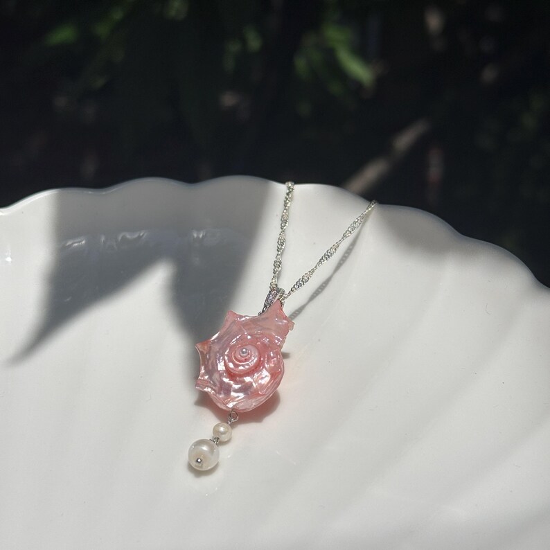 Puede incluir: Un collar con un colgante de concha marina rosa pulida con un dise&ntilde;o en espiral, suspendido de una cadena de plata. Dos peque&ntilde;as perlas blancas redondas est&aacute;n unidas a la parte inferior de la concha, creando un aspecto delicado.