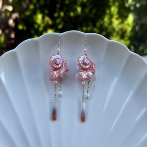 Puede incluir: Un par de pendientes colgantes con dijes de concha espiral rosa iridiscente suspendidos de ganchos plateados. Cada pendiente incluye peque&ntilde;as perlas blancas, cadenas plateadas delicadas y un colgante de gota delgado con degradado en blanco y naranja.