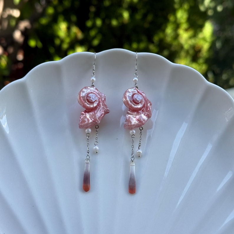 Puede incluir: Un par de pendientes colgantes con dijes de concha espiral rosa iridiscente suspendidos de ganchos plateados. Cada pendiente incluye peque&ntilde;as perlas blancas, cadenas plateadas delicadas y un colgante de gota delgado con degradado en blanco y naranja.