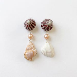 May include: Dangle earrings with a red and white patterned bead, a peach pearl, and a seashell. One earring has a tan seashell, the other a white one. The earrings are set against a white backdrop.