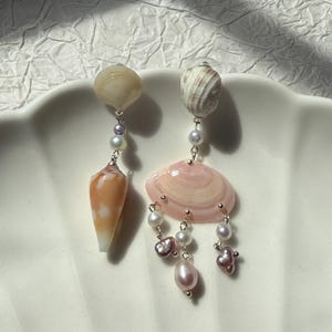 May include: A pair of dangle earrings featuring natural seashells and freshwater pearls. One earring has a light orange conch shell, while the other has a pink shell. Both earrings have pearl accents and silver-tone metal findings.