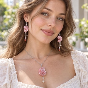 Puede incluir: Un conjunto de joyer&iacute;a compuesto por un collar y pendientes colgantes a juego. Cada pieza presenta un colgante de concha espiral rosa, acentuado con peque&ntilde;as perlas blancas y un dije transl&uacute;cido y delgado en forma de l&aacute;grima de color rojo rosado.
