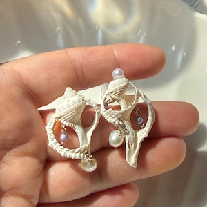 May include: A pair of seashell earrings. The earrings are white with intricate shell detailing. Each earring features small, dangling pearls and lavender beads. The earrings are held in a hand.