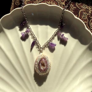 May include: A silver-toned chain necklace featuring a central teardrop-shaped pendant with a textured white border and a light purple geode center. The chain is accented with several small, irregular, polished purple amethyst stone beads.