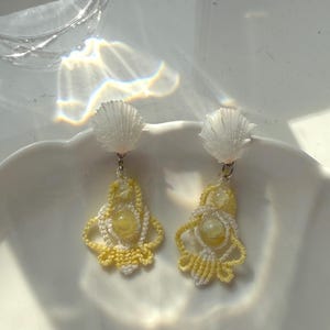May include: A pair of drop earrings featuring white seashell tops attached to intricate, yellow and white woven thread pendants. Each pendant contains a central, smooth, pale yellow bead surrounded by delicate, knotted fiber patterns.