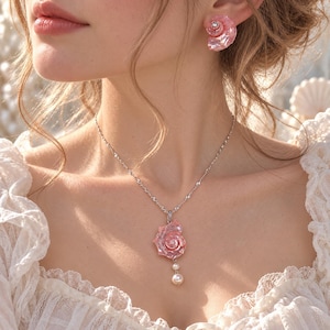 Puede incluir: Un conjunto de joyer&iacute;a que consiste en un collar de cadena plateada con un colgante de concha marina rosa iridiscente acentuado por dos peque&ntilde;as perlas blancas, y un pendiente de concha a juego con una piedra de cristal transparente.