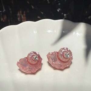 Puede incluir: Un par de pendientes de bot&oacute;n con forma de peque&ntilde;as conchas marinas de color rosa iridiscente. Cada concha presenta un acabado brillante y semitransl&uacute;cido con un cristal transparente facetado incrustado en el centro de la espiral.