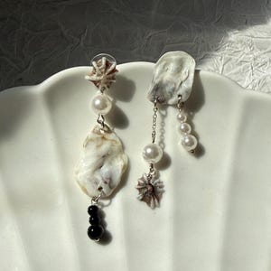May include: Asymmetrical earrings with natural seashells, pearls, and dark beads. One earring has a seashell with a pearl and black beads. The other has a seashell, pearls, and a small chain.