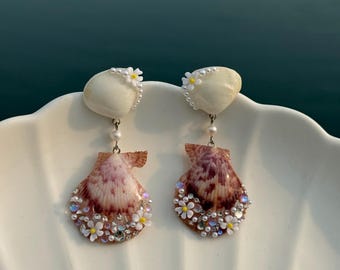 以下が含まれることがあります： 小さな白い花が添えられた滑らかな白い貝殻のトップと、赤茶色の模様が入った扇形の貝殻が揺れるピアス。小さな白い花、透明なクリスタル、そして小さな白いビーズで装飾されています。