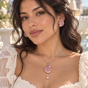 Puede incluir: Un conjunto de joyer&iacute;a que consiste en un collar de cadena plateada con un colgante de concha marina rosa y una perla blanca colgante, junto con pendientes de bot&oacute;n a juego en forma de concha rosa. El collar presenta una cadena de eslabones.