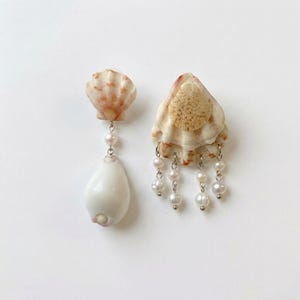 May include: A pair of seashell earrings. One earring features a small, light-colored shell with a dangling white teardrop-shaped shell and pearls. The other earring has a larger shell with dangling pearls.